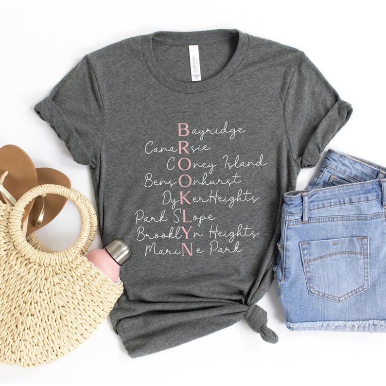 May include: A heather gray t-shirt with a white graphic that says "BROOKLYN" with the letters spelled out vertically using the names of different neighborhoods in Brooklyn, New York. The letters are in pink.