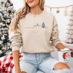 Op de afbeelding: Beige crewneck sweatshirt met een minimalistisch winterontwerp. De afbeelding toont drie sneeuwmannen, een kerstboom en een dunne zwarte lijn. De sweatshirt is gemaakt van een zacht materiaal en is geschikt voor casual wear.
