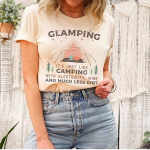 May include: A cream-coloured t-shirt with the text "GLAMPING" in black letters. The text "IT'S JUST LIKE CAMPING WITH ELECTRICITY, WINE AND MUCH LESS DIRT" is printed below the word "GLAMPING". A cartoon image of a tent with string lights is in the centre of the shirt.