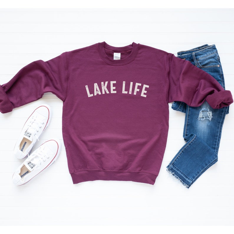 May include: A maroon sweatshirt with the text "LAKE LIFE" in white on the front. The sweatshirt is laid flat on a white surface with a pair of white sneakers and blue jeans.