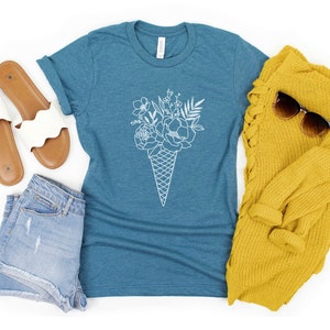 May include: A teal blue t-shirt featuring a white outline of a bouquet of flowers in an ice cream cone. The t-shirt is laid out on a white surface with a pair of white sandals, denim shorts, and a yellow sweater.