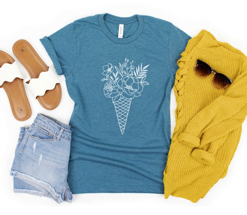 May include: A teal blue t-shirt featuring a white outline of a bouquet of flowers in an ice cream cone. The t-shirt is laid out on a white surface with a pair of white sandals, denim shorts, and a yellow sweater.