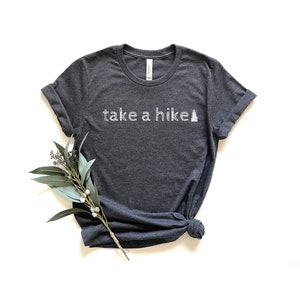 May include: A dark heather gray t-shirt with the text "take a hike" and a stylized pine tree graphic in white.