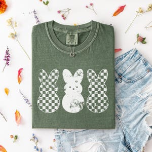 May include: Olive green t-shirt featuring three white bunny designs. Two bunnies have a checkered pattern, and one has a solid design. The shirt is paired with denim shorts and floral accents, creating a casual, spring-themed aesthetic.