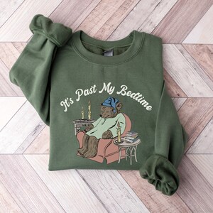 Pu&ograve; includere: Una felpa verde con il testo "It's Past My Bedtime" in bianco. Un orso dei cartoni animati con un berretto da notte blu &egrave; seduto su una sedia con un libro e una candela accesa.