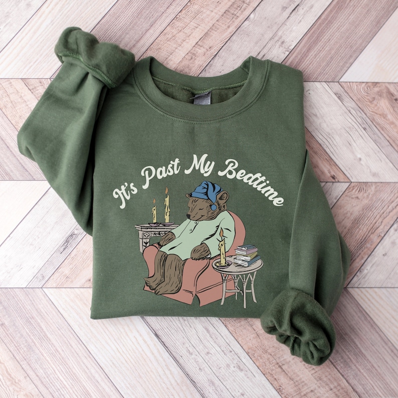 Pu&ograve; includere: Una felpa verde con il testo "It's Past My Bedtime" in bianco. Un orso dei cartoni animati con un berretto da notte blu &egrave; seduto su una sedia con un libro e una candela accesa.