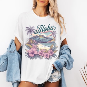 以下が含まれることがあります： 白いTシャツ。ヤシの木、山、ビーチなど、熱帯の風景が描かれています。「Aloha」の文字はターコイズ色です。ピンクのハイビスカスの花もデザインされています。ゆったりとしたフィット感のTシャツです。