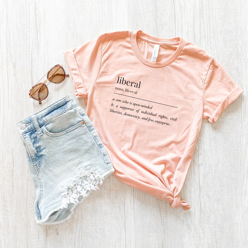 May include: A peach-colored t-shirt with the word "liberal" defined as "one who is open-minded" and "a supporter of individual rights, civil liberties, democracy, and free enterprise."
