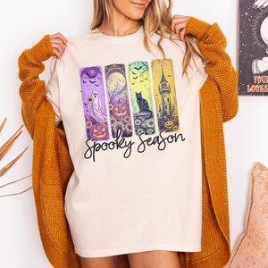 May include: Cream-colored t-shirt with a "Spooky Season" graphic. The graphic features four vertical panels with Halloween-themed illustrations: a ghost, pumpkins, a black cat, and a haunted tower. The shirt is paired with a mustard-colored cardigan.