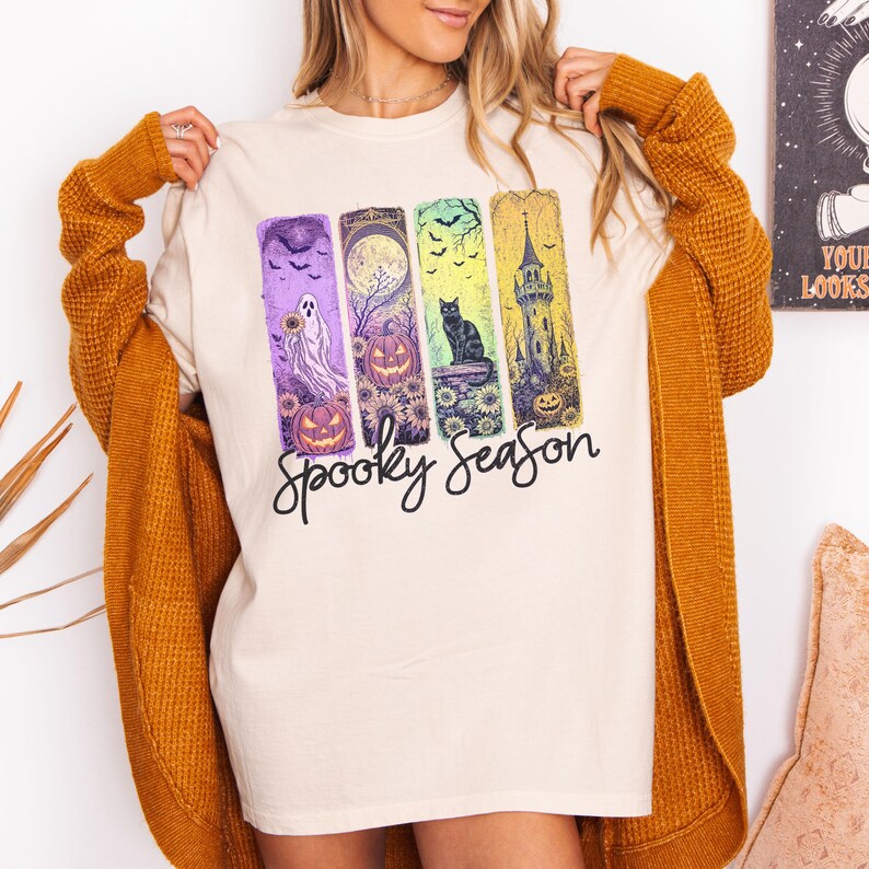 May include: Cream-colored t-shirt with a "Spooky Season" graphic. The graphic features four vertical panels with Halloween-themed illustrations: a ghost, pumpkins, a black cat, and a haunted tower. The shirt is paired with a mustard-colored cardigan.