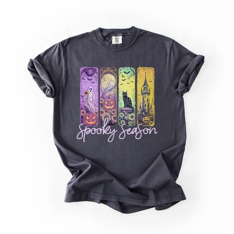 May include: Dark gray t-shirt featuring a colorful Halloween-themed design. The design includes a ghost, pumpkins, a black cat, and a spooky tower. The words "Spooky Season" are written in a cursive font below the design.