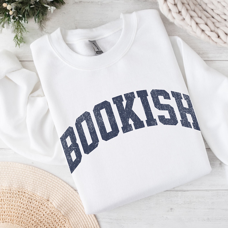 May include: White crewneck sweatshirt with the word "BOOKISH" printed in a navy blue, distressed, collegiate-style font.