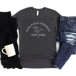 Puede incluir: Una camiseta gris oscuro con un dise&ntilde;o gr&aacute;fico blanco de un caballo y un jinete. El gr&aacute;fico incluye el texto "Saratoga Springs New York 1819".