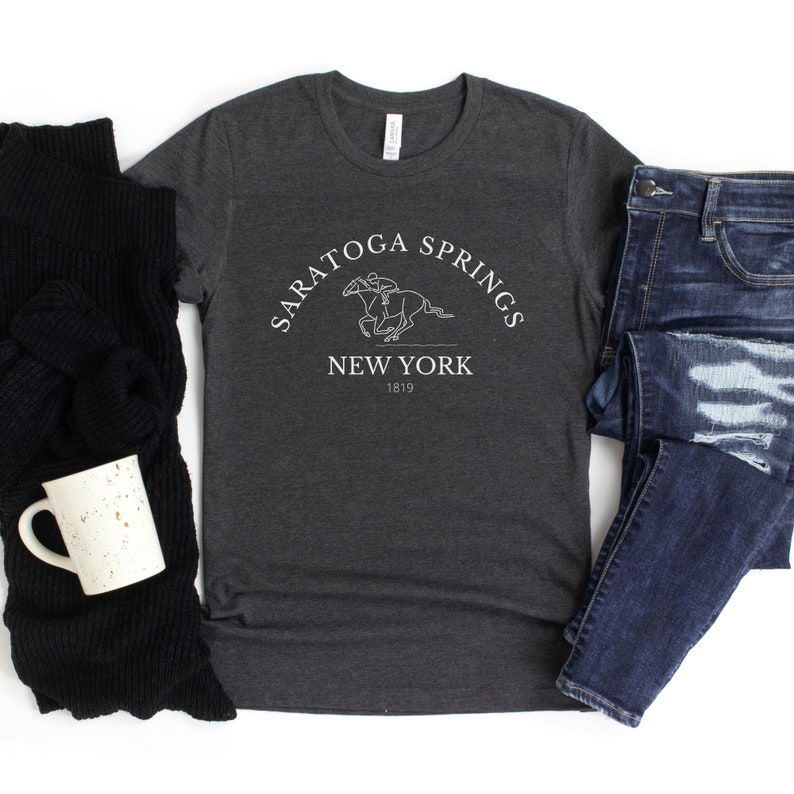 Puede incluir: Una camiseta gris oscuro con un dise&ntilde;o gr&aacute;fico blanco de un caballo y un jinete. El gr&aacute;fico incluye el texto "Saratoga Springs New York 1819".