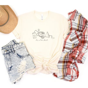 May include: A beige t-shirt with a graphic of tea bags, a teacup, and a tea bag tag. The text "it's a tea shirt" is printed below the graphic.