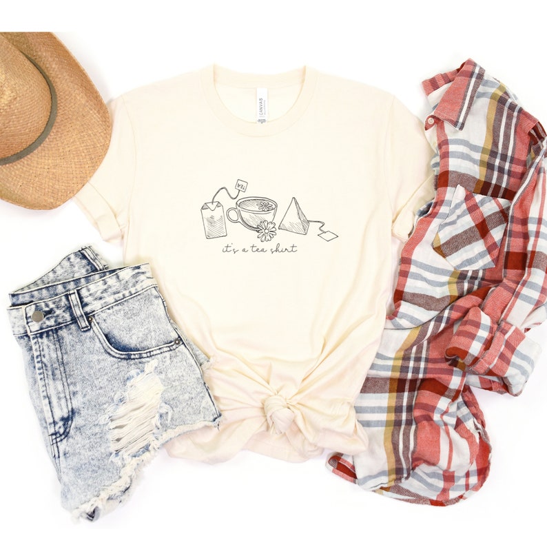 May include: A beige t-shirt with a graphic of tea bags, a teacup, and a tea bag tag. The text "it's a tea shirt" is printed below the graphic.