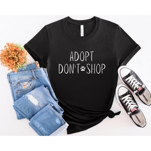 May include: Black t-shirt with white text that reads "Adopt Don't Shop" and a paw print. The shirt is laying on a white surface with a pair of blue jeans and black sneakers.