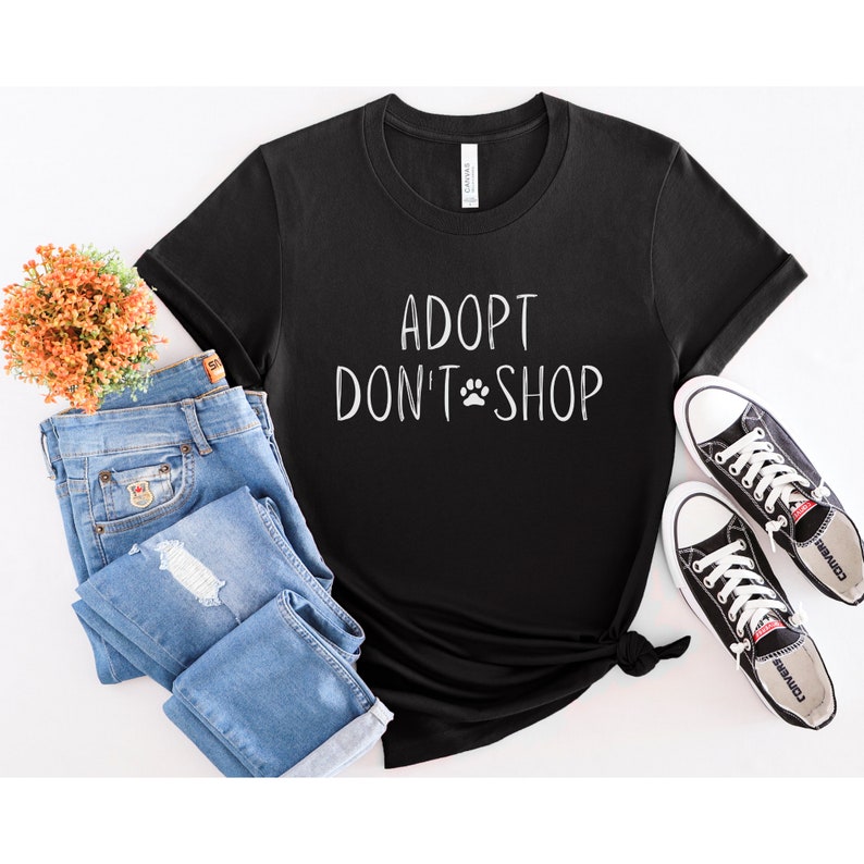May include: Black t-shirt with white text that reads "Adopt Don't Shop" and a paw print. The shirt is laying on a white surface with a pair of blue jeans and black sneakers.