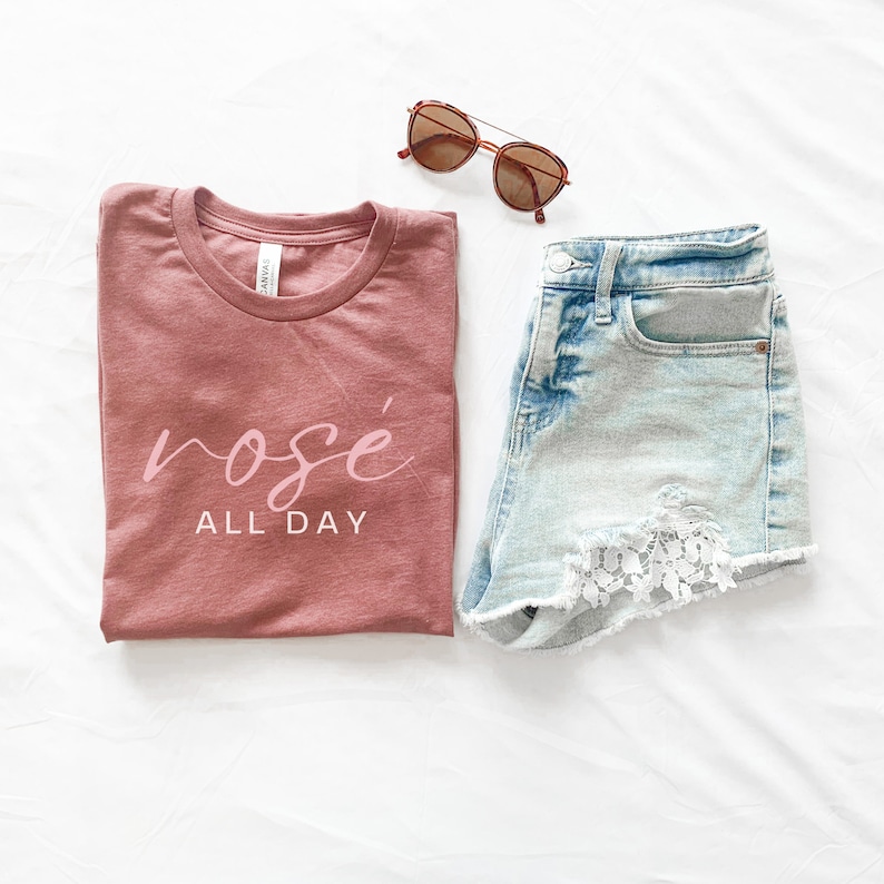 May include: A folded mauve t-shirt with the text "ros&eacute; all day" in pink lettering. A pair of light blue denim shorts with white lace trim and brown sunglasses are also pictured.