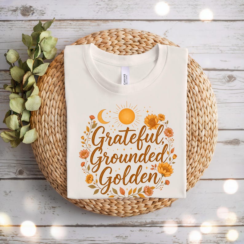 Pu&ograve; includere: T-shirt color crema con le parole "Grateful, Grounded, Golden" in un carattere caldo autunnale. Il design include un sole, una luna ed elementi floreali nei toni dell'arancione e del marrone. La maglietta &egrave; esposta su un cesto intrecciato.