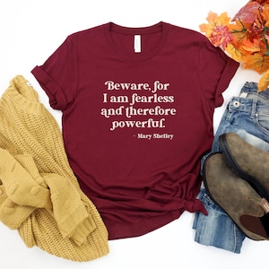 Puede incluir: Camiseta color burdeos con texto blanco que dice "Beware, for I am fearless and therefore powerful. - Mary Shelley". La camiseta est&aacute; colocada sobre una superficie blanca con un su&eacute;ter amarillo, jeans azules y botas marrones.