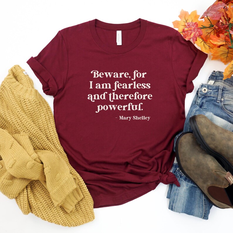 Puede incluir: Camiseta color burdeos con texto blanco que dice "Beware, for I am fearless and therefore powerful. - Mary Shelley". La camiseta est&aacute; colocada sobre una superficie blanca con un su&eacute;ter amarillo, jeans azules y botas marrones.