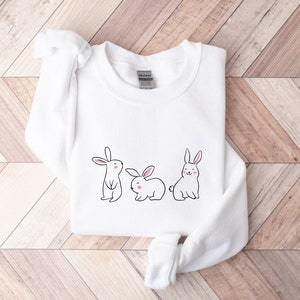 May include: White sweatshirt featuring a design of three cartoon bunnies. The bunnies are white with pink cheeks and black outlines.