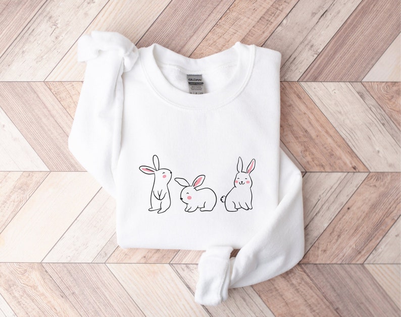 May include: White sweatshirt featuring a design of three cartoon bunnies. The bunnies are white with pink cheeks and black outlines.