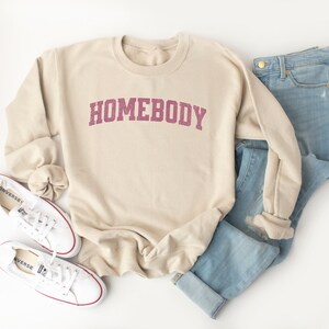 Puede incluir: Una sudadera beige con la palabra "HOMEBODY" impresa en color granate en la parte delantera. La sudadera est&aacute; colocada sobre una superficie blanca con un par de zapatillas blancas y unos vaqueros azules.