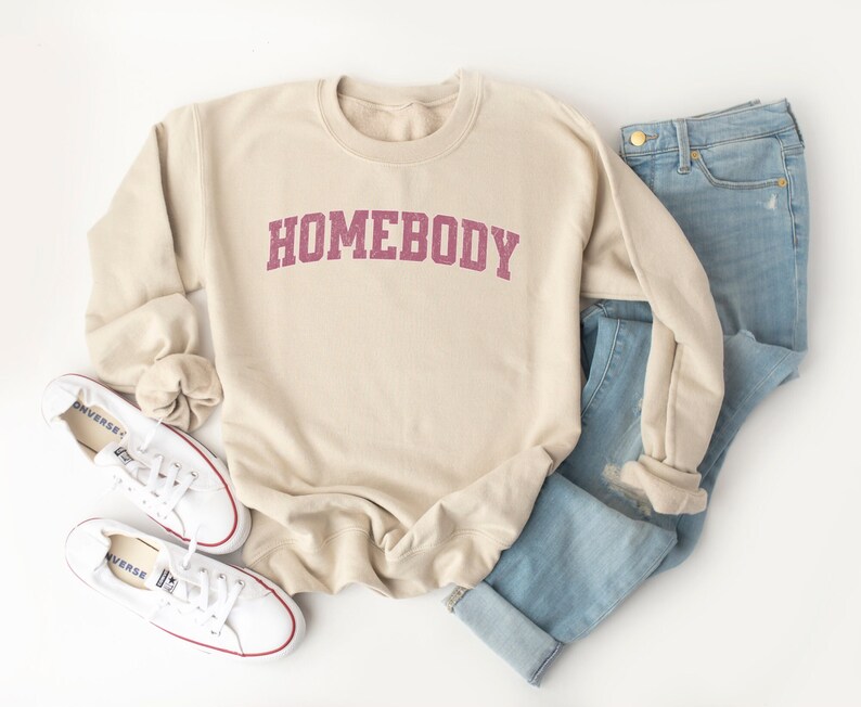 Puede incluir: Una sudadera beige con la palabra "HOMEBODY" impresa en color granate en la parte delantera. La sudadera est&aacute; colocada sobre una superficie blanca con un par de zapatillas blancas y unos vaqueros azules.
