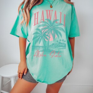 May include: A turquoise blue t-shirt with a graphic print of palm trees and a sunset. The text "Hawaii Aloha State" is printed in pink on the shirt.