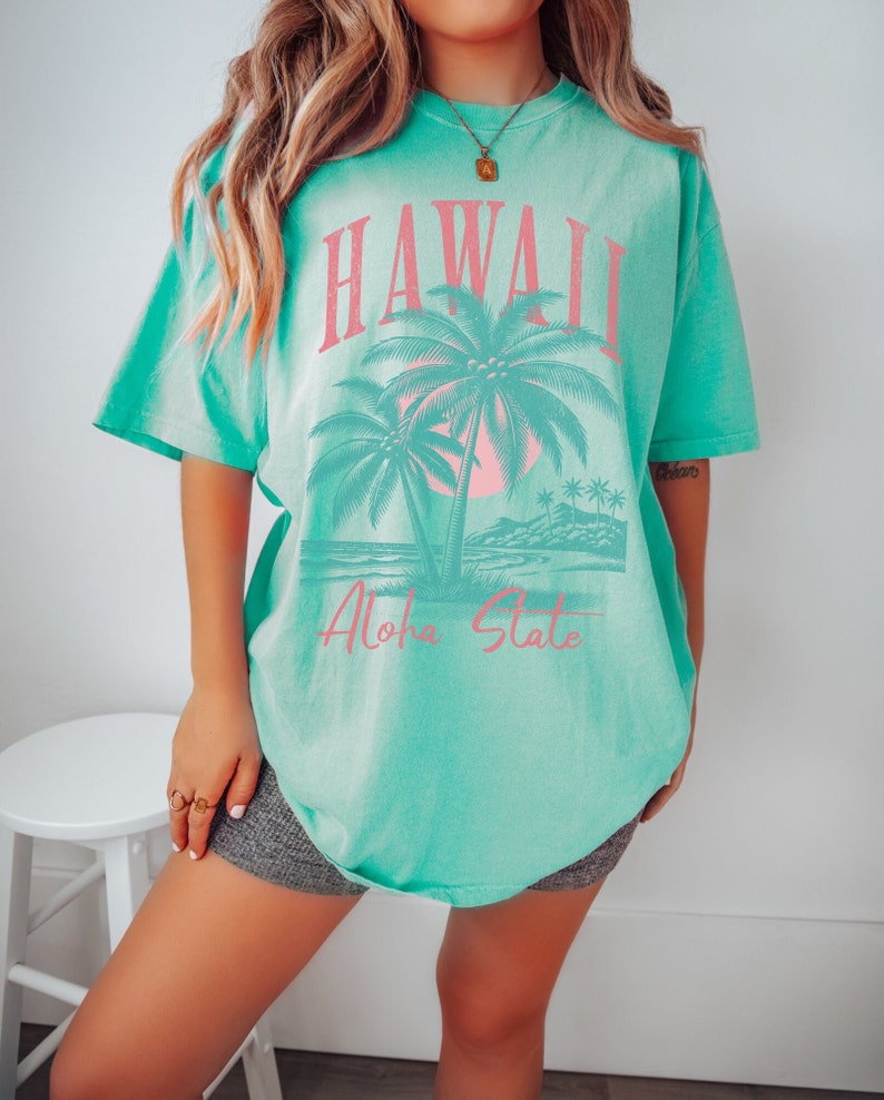 May include: A turquoise blue t-shirt with a graphic print of palm trees and a sunset. The text "Hawaii Aloha State" is printed in pink on the shirt.
