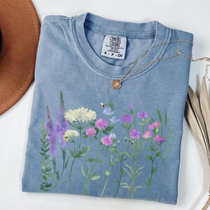 May include: A blue Comfort Colors t-shirt featuring a floral design with purple, pink, and white flowers, and a small bee. A gold necklace with a pendant rests on the shirt. The shirt has a tag that says "Comfort Colors".