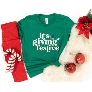 May include: A green t-shirt with the text "it's giving festive" in white. Red leggings and a candy cane are on the left. A white faux fur stocking with red ornaments and a poinsettia are on the right. Festive holiday apparel.