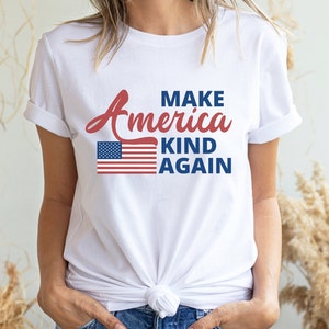 May include: White t-shirt with a red, white, and blue American flag graphic and the text "Make America Kind Again".