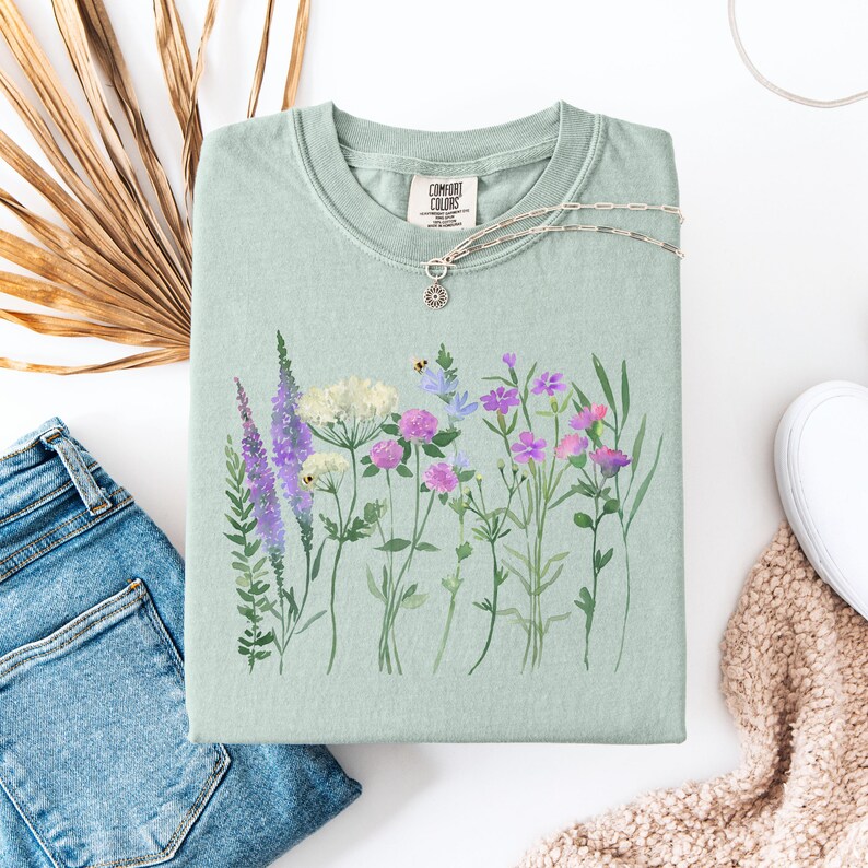 May include: A light green Comfort Colors sweatshirt featuring a watercolor floral design with purple, pink, and white flowers. The shirt is paired with blue jeans, a silver necklace, and a white sneaker.