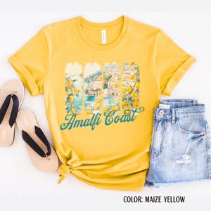 May include: Maize yellow t-shirt featuring an Amalfi Coast graphic. The design shows a scenic view with lemons and teal text reading "Amalfi Coast". The shirt is styled with denim shorts and sandals.