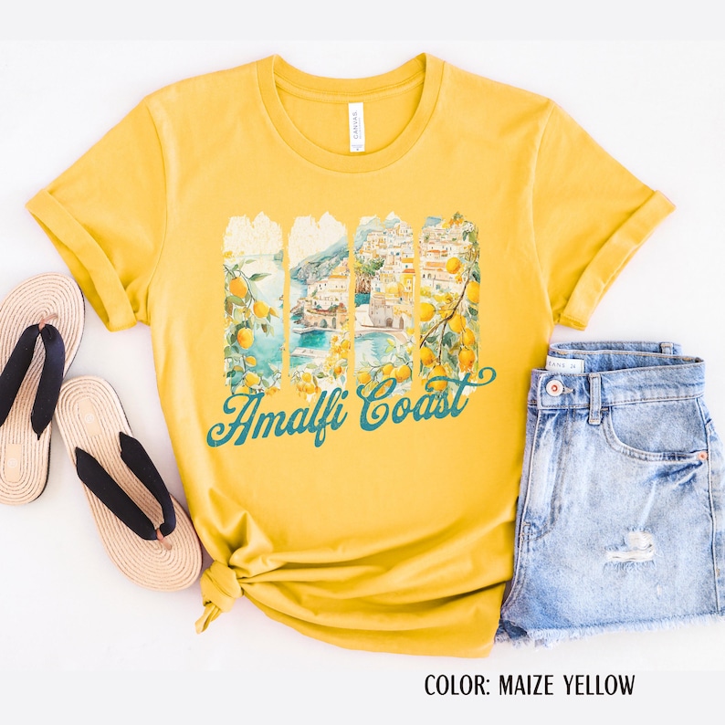 May include: Maize yellow t-shirt featuring an Amalfi Coast graphic. The design shows a scenic view with lemons and teal text reading "Amalfi Coast". The shirt is styled with denim shorts and sandals.