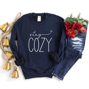 May include: A navy blue sweatshirt with the text "stay cozy" in white lettering. The sweatshirt is laid flat on a white background with a pair of blue jeans and a sprig of red poinsettia.