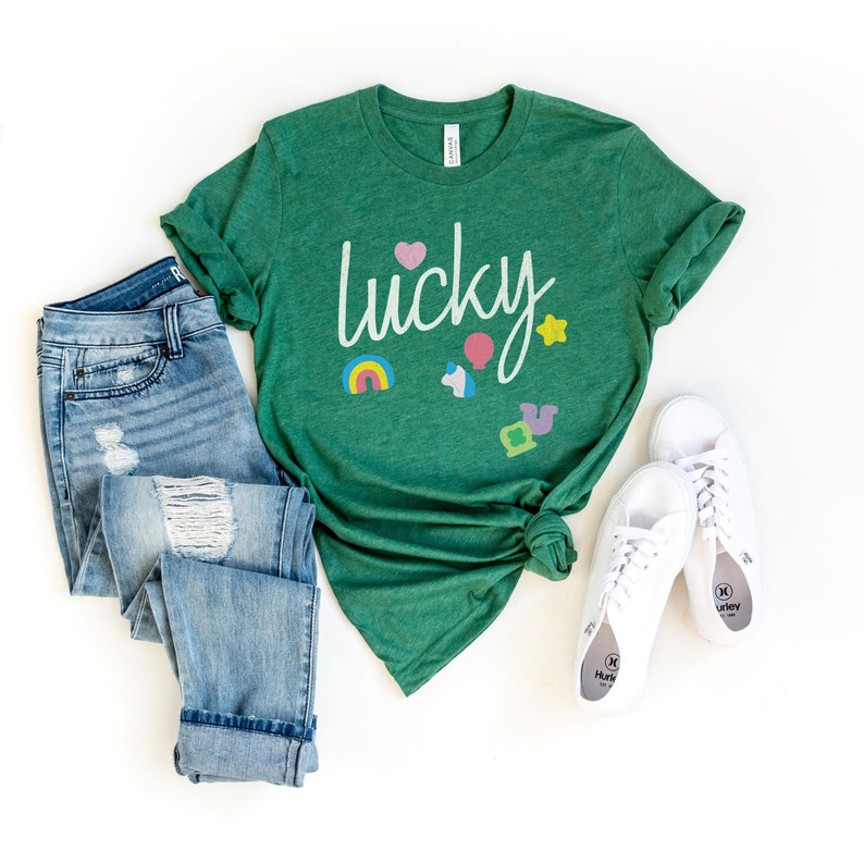 Puede incluir: Una camiseta verde con la palabra "lucky" escrita en blanco. La camiseta tiene un arco&iacute;ris, una herradura, un tr&eacute;bol de cuatro hojas, un unicornio y un coraz&oacute;n impreso en ella.