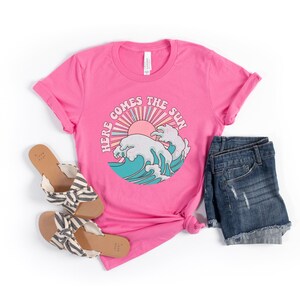 Op de afbeelding: Roze T-shirt met een afbeelding van een zon en golven met de tekst "Here Comes The Sun". Het shirt wordt gecombineerd met een spijkerbroekje en sandalen.