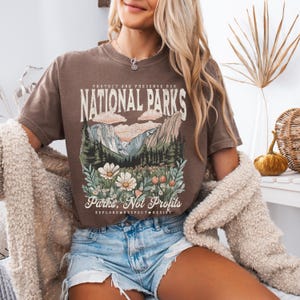 K&ouml;nnte beinhalten: Ein braunes T-Shirt mit einem grafischen Aufdruck einer Berglandschaft und dem Text "Sch&uuml;tzen und bewahren Sie unsere Nationalparks" und "Parks, nicht Profite" mit den Worten "Erkunden, Respektieren, Widerstehen" darunter.