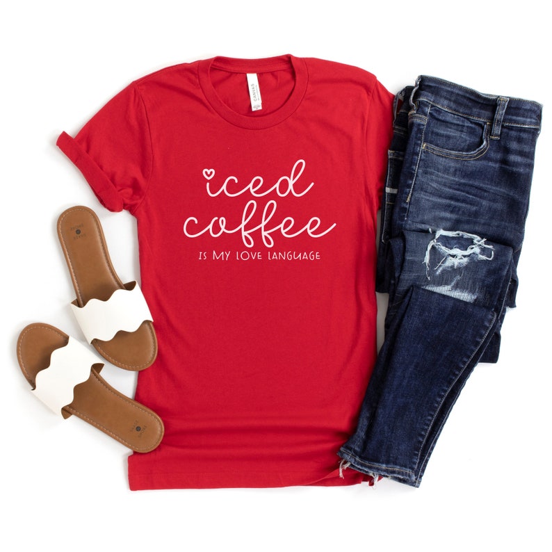 Puede incluir: Camiseta roja con texto blanco que dice "iced coffee is my love language". La camiseta se combina con jeans azules y sandalias marrones.