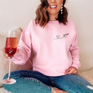 Op de afbeelding: Een roze sweatshirt met een kleine zwarte afbeelding van een papieren vliegtuig met een hartje aan de staart. Het sweatshirt wordt gedragen door iemand die een glas champagne vasthoudt.