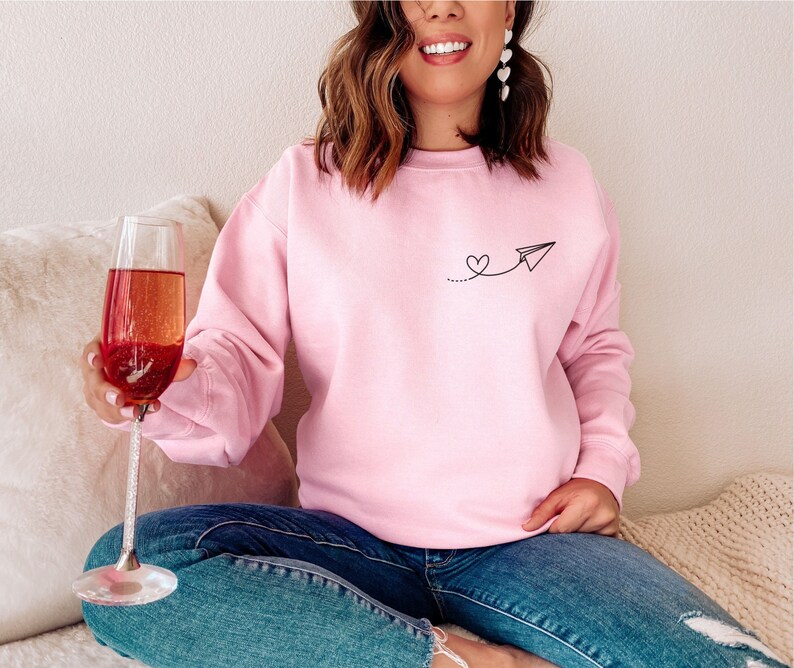 Op de afbeelding: Een roze sweatshirt met een kleine zwarte afbeelding van een papieren vliegtuig met een hartje aan de staart. Het sweatshirt wordt gedragen door iemand die een glas champagne vasthoudt.