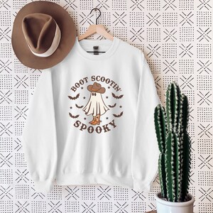 Op de afbeelding: Wit sweatshirt met een bruine afbeelding van een spook dat een cowboyhoed en laarzen draagt. De tekst "Boot Scootin' Spooky" staat op het sweatshirt gedrukt.