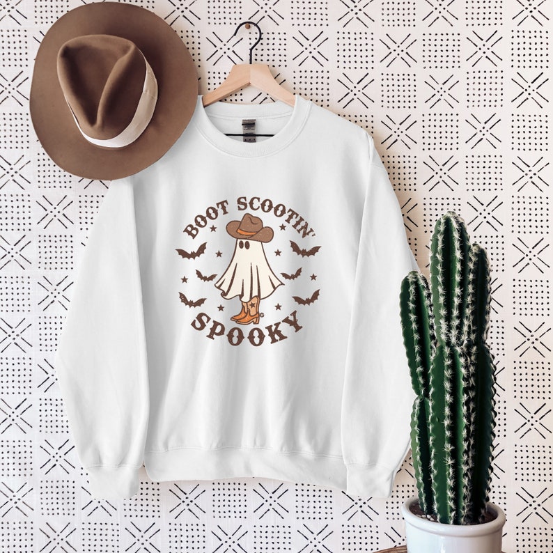 Op de afbeelding: Wit sweatshirt met een bruine afbeelding van een spook dat een cowboyhoed en laarzen draagt. De tekst "Boot Scootin' Spooky" staat op het sweatshirt gedrukt.