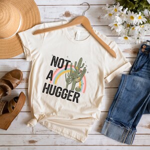 May include: A beige t-shirt with a graphic of a cactus and a rainbow with the text "NOT A HUGGER" in black.