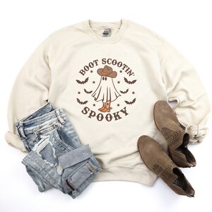 Op de afbeelding: Een beige sweatshirt met een afbeelding van een spook dat een cowboyhoed en laarzen draagt. De tekst "Boot Scootin' Spooky" staat op het sweatshirt gedrukt.