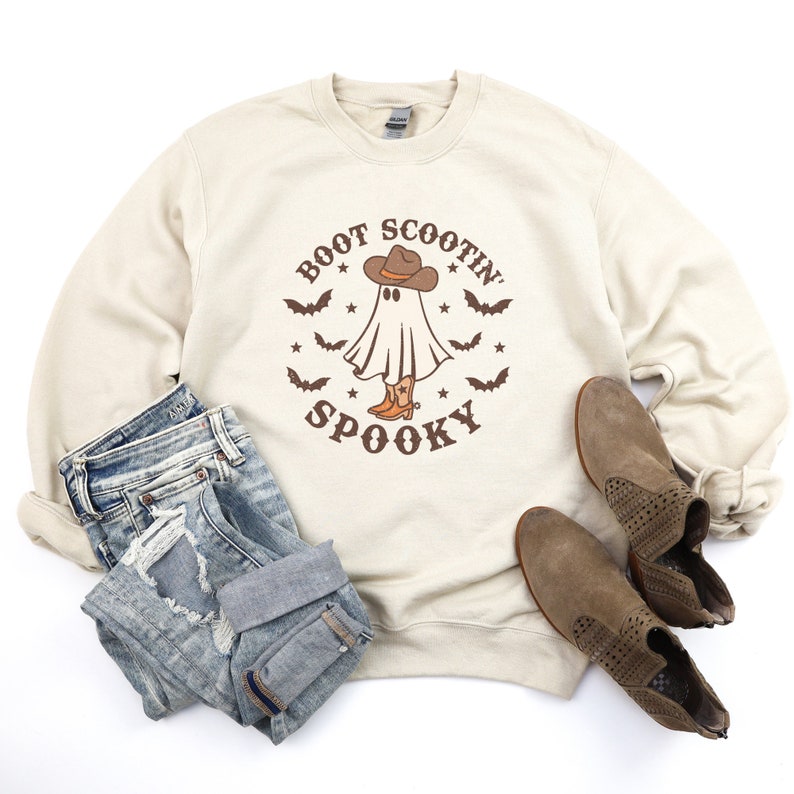 Op de afbeelding: Een beige sweatshirt met een afbeelding van een spook dat een cowboyhoed en laarzen draagt. De tekst "Boot Scootin' Spooky" staat op het sweatshirt gedrukt.
